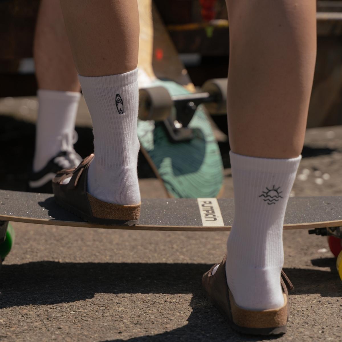 Person standing on a surfskate wearing white Synix SunSeeker sustainable socks focus on surfboard